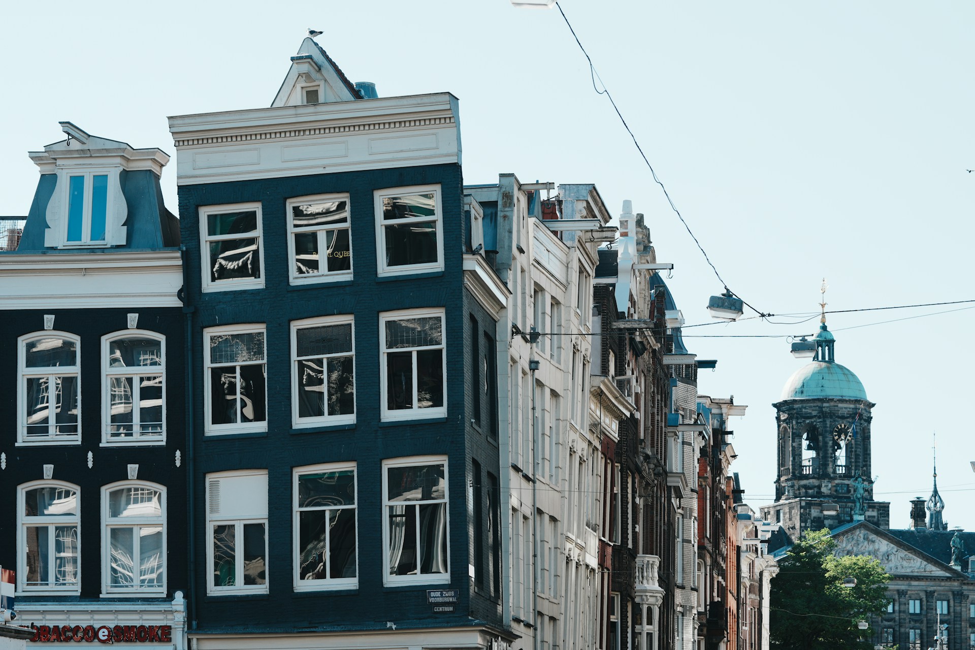 Street view of traditional narrow buildings with large windows and a clock tower with a green dome in the background.