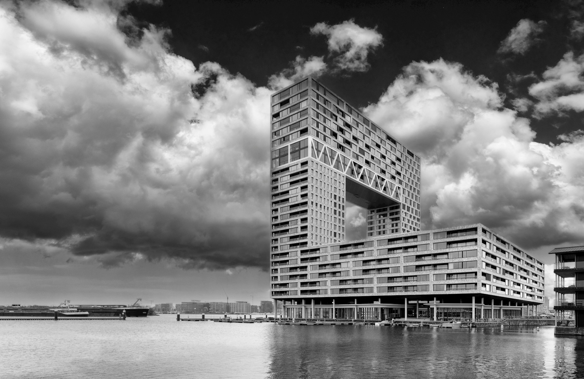 Modern architectural building with a large rectangular cutout over calm water under a dramatic cloudy sky.